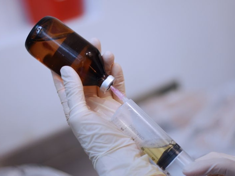 mano enguantada extrayendo líquido de un vial farmacéutico ámbar con una jeringa, sobre un fondo claro.